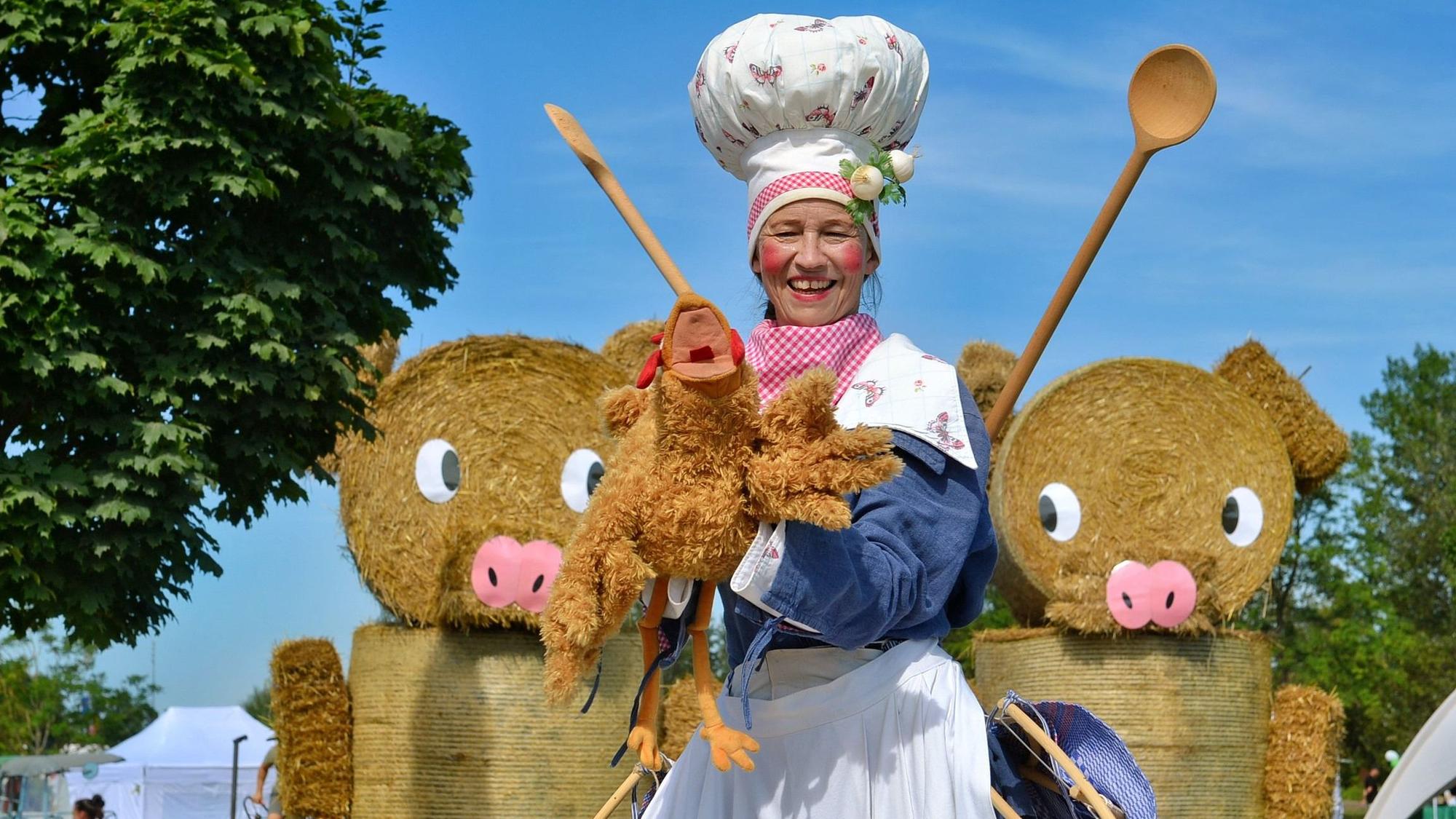 Eine Frau mit großer weiß-roter Kochmütze, zwei Kochlöffeln auf dem Rücken und weißer Schürze hält ein Stoffhuhn in die Kamera und lacht. Im Hintergrund sind zwei große Schweine-Figuren aus Strohballen zu erkennen.