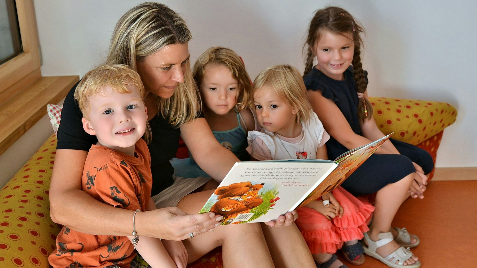 Vier Kinder sitzen zusammen mit einer blonden Frau auf einem gelb-roten Sofa und schauen sich ein Buch an.