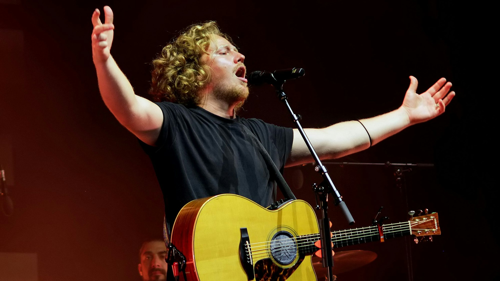 Das Bild zeigt den Sänger, er breitet die Arme aus und singt in ein Mikrofon.