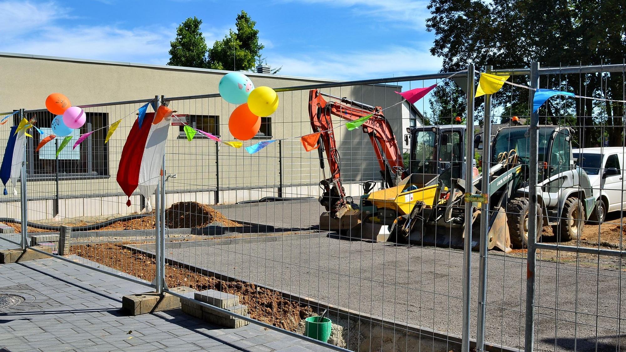 An einem Bauzaun, der vor zwei Baggern steht, hängen bunte Luftballons.