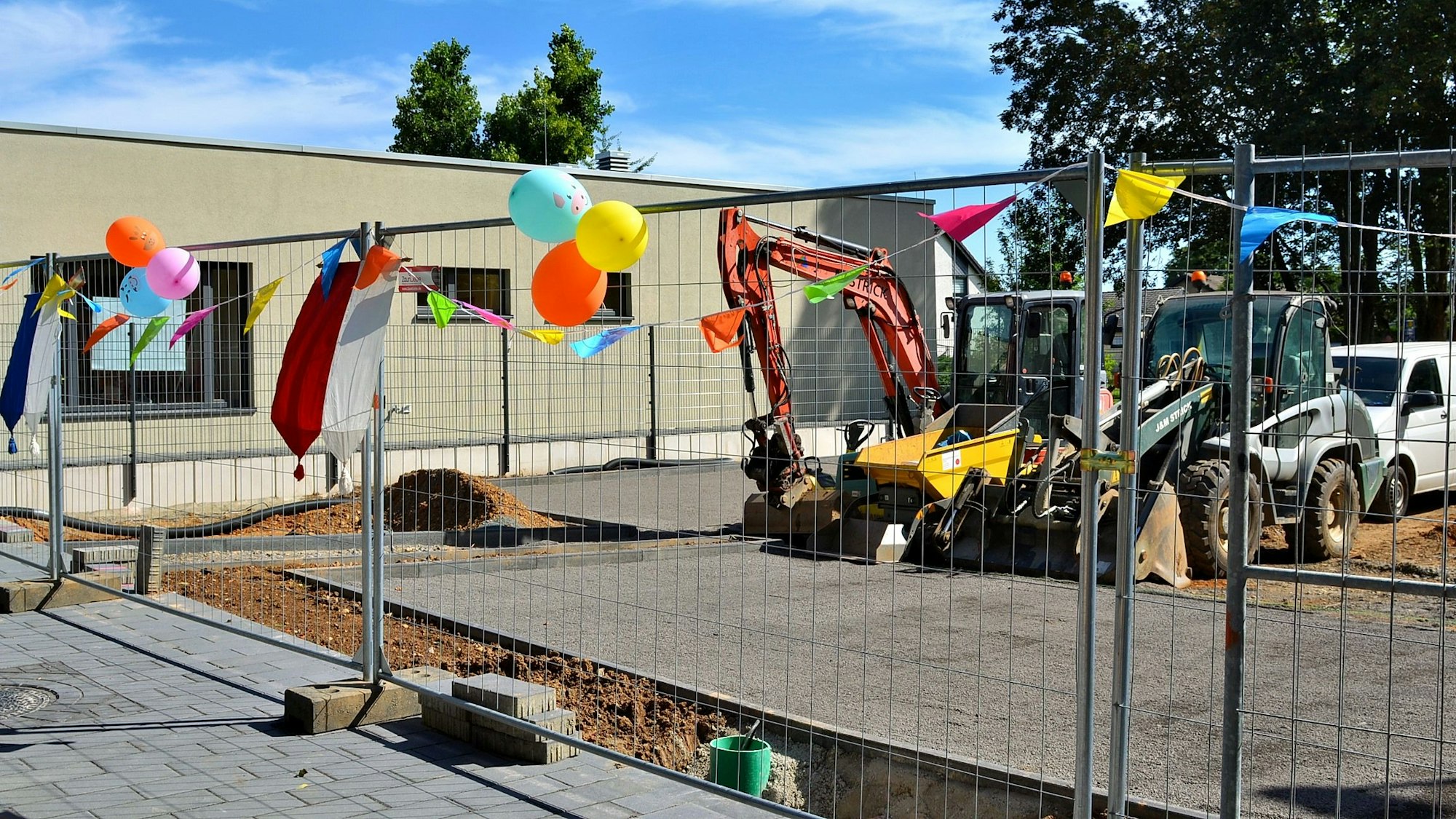 An einem Bauzaun, der vor zwei Baggern steht, hängen bunte Luftballons.