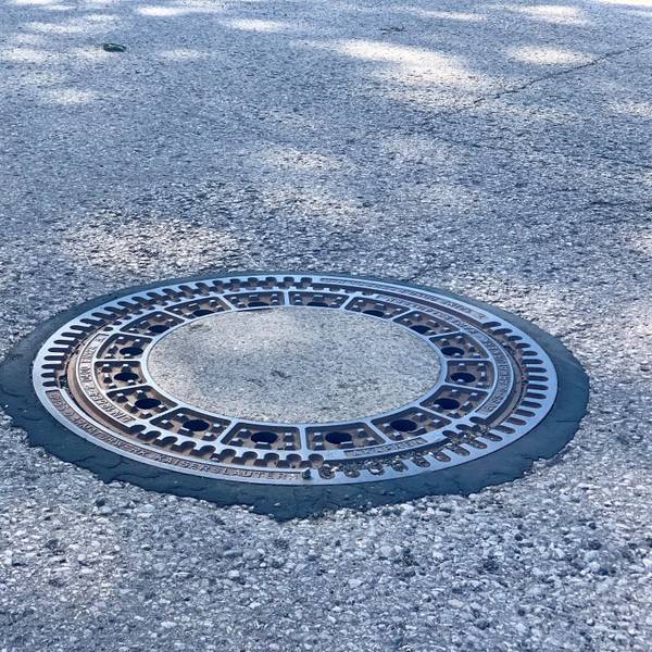 Zu sehen ist ein runder Gulli in einer Straße.