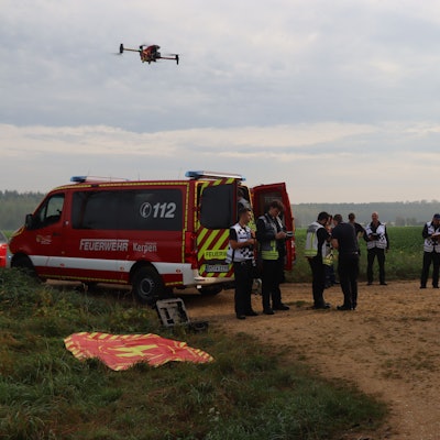 Feuerwehrleute stehen an einem Einsatzfahrzeug, am Himmel fliegt eine Drohne.