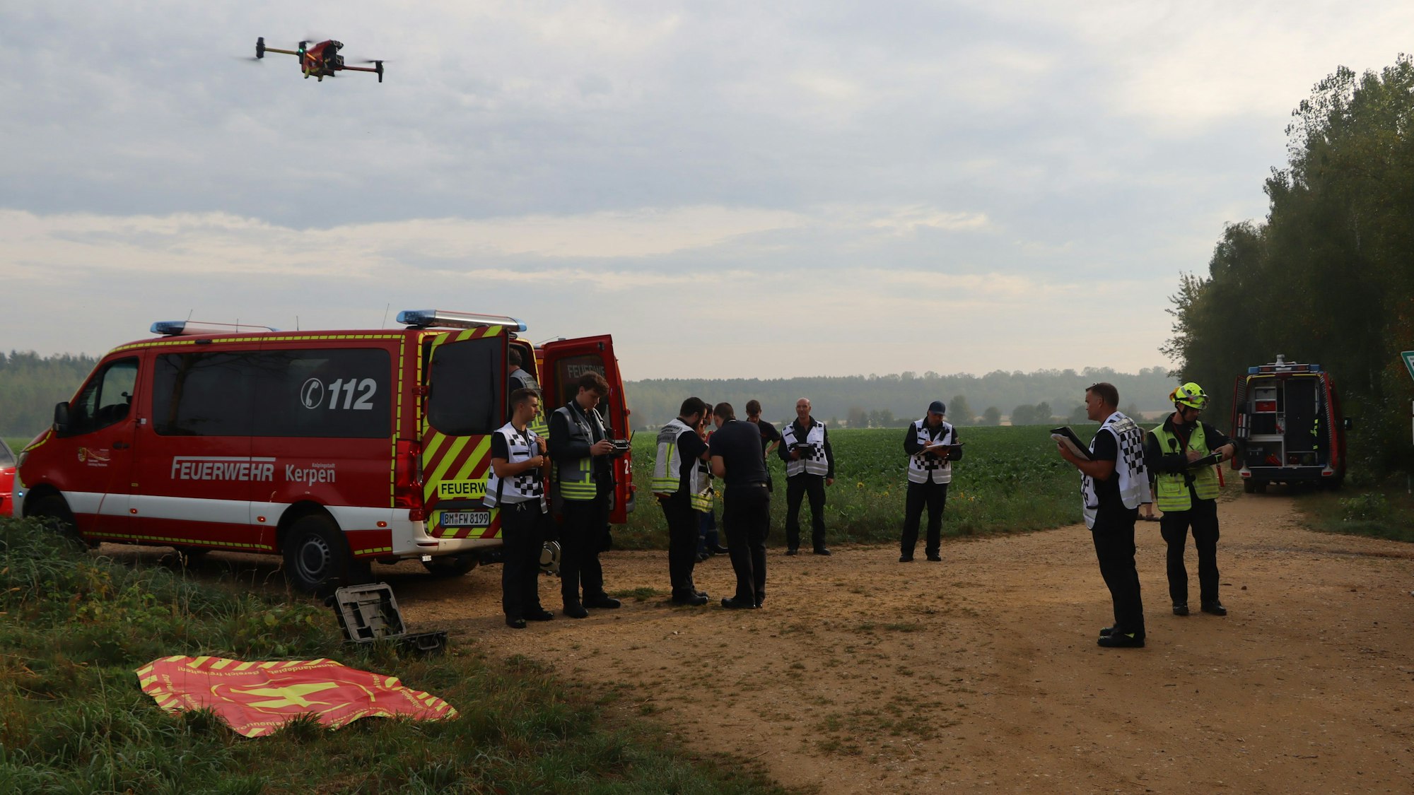 Feuerwehrleute stehen an einem Einsatzfahrzeug, am Himmel fliegt eine Drohne.