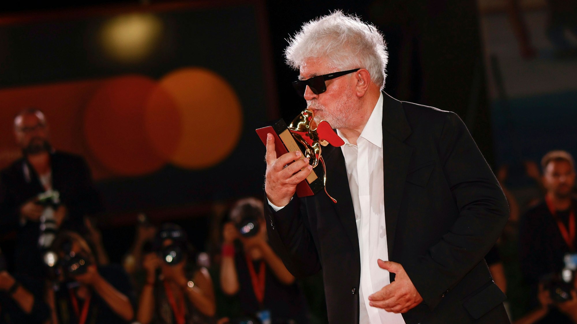 Pedro Almodóvar steht in einem schwarzen Anzug vor Fotografen und küsst den Goldenen Löwen, den er gewonnen hat