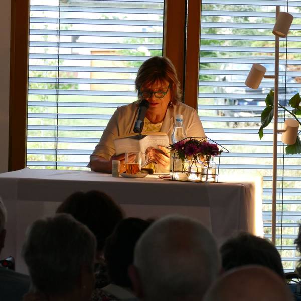 Eine Frau mit Brille sitzt an einem Tisch mit einer Leselampe und liest aus einem Buch vor.