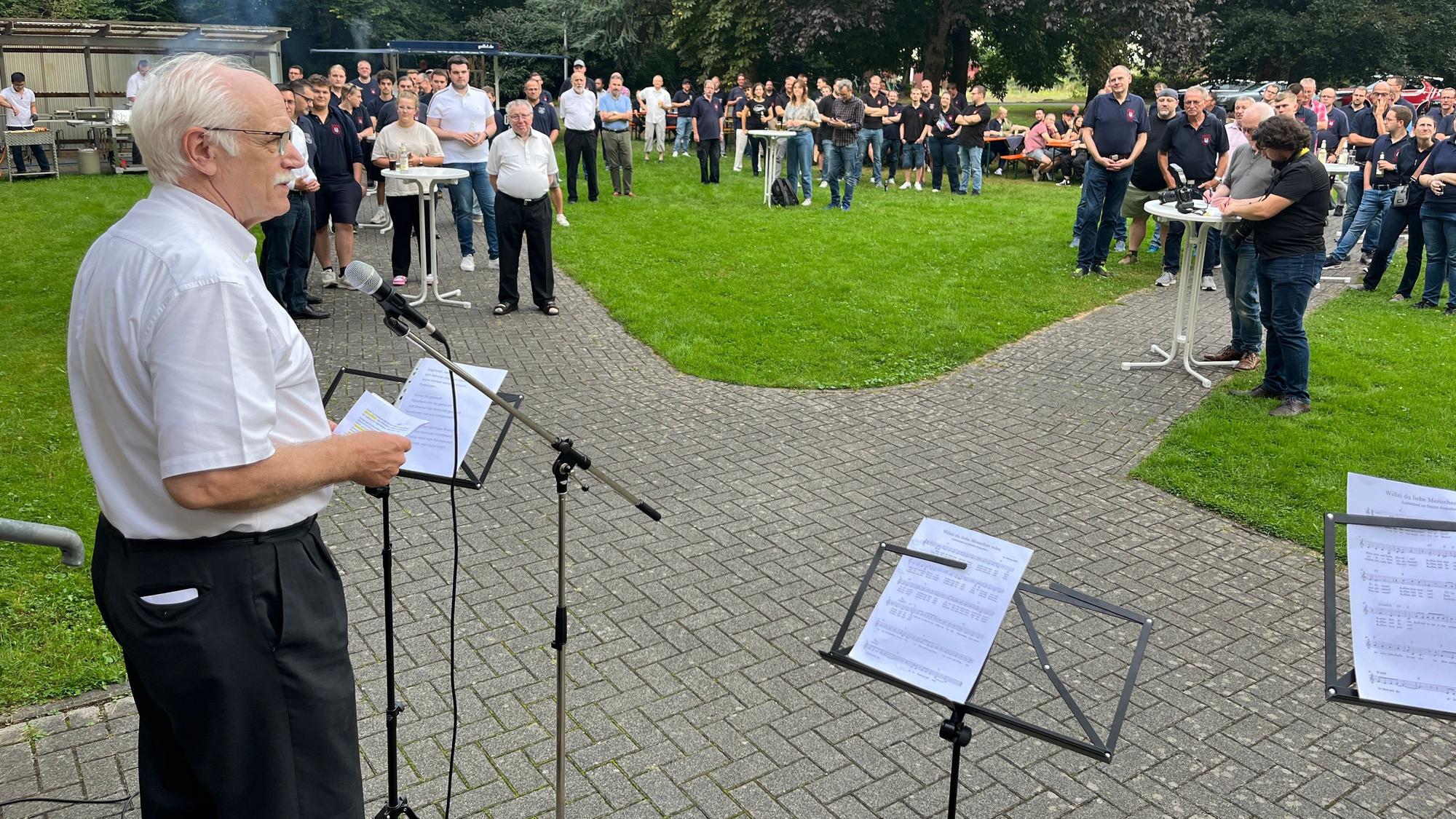 Ein Pater steht vor Feuerwehrleuten und hält eine Rede.