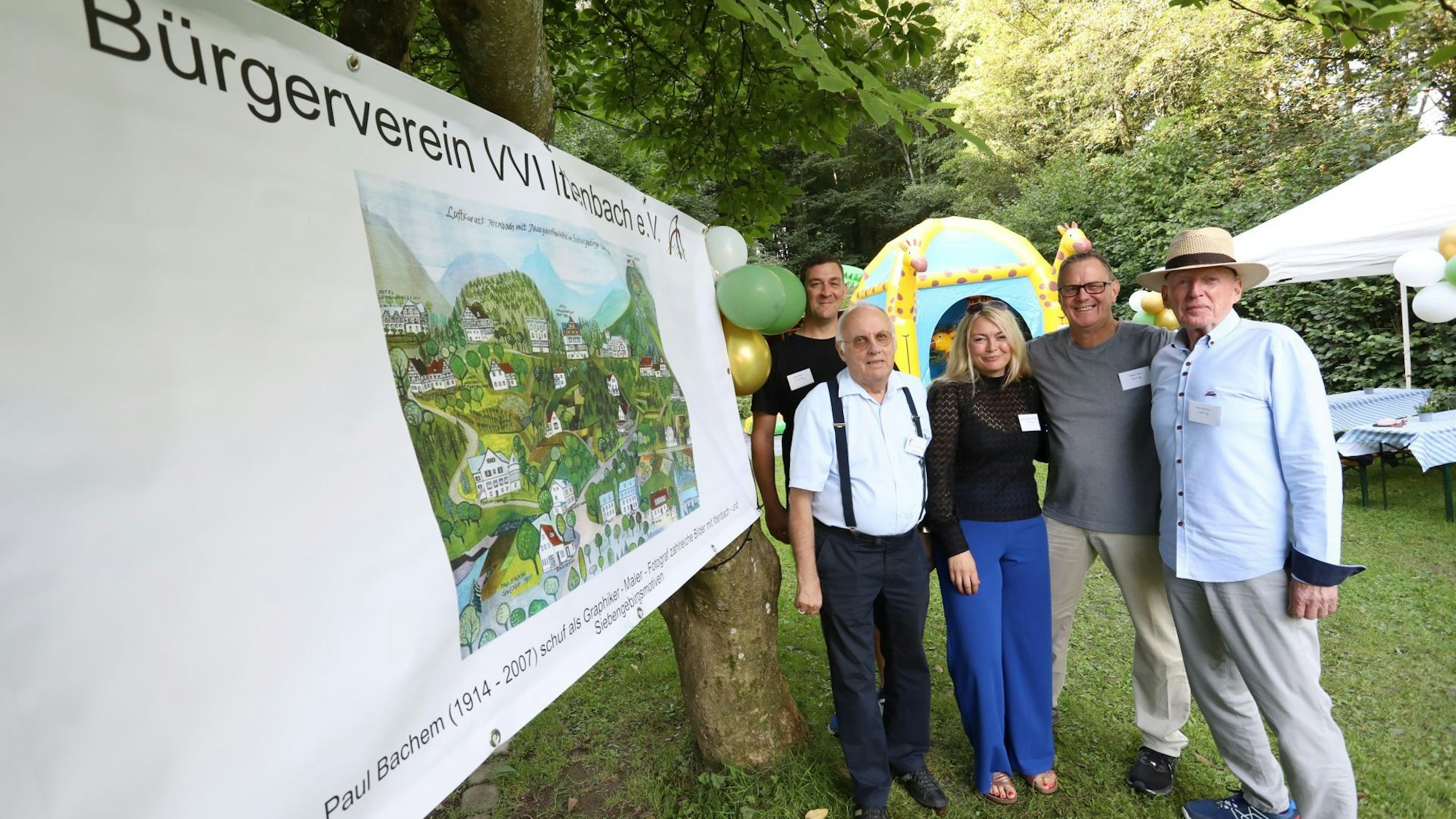 Männer und eine Frau stehen auf einer Wiese, auf der Zelte und Sonnenschirme zu sehen sind.