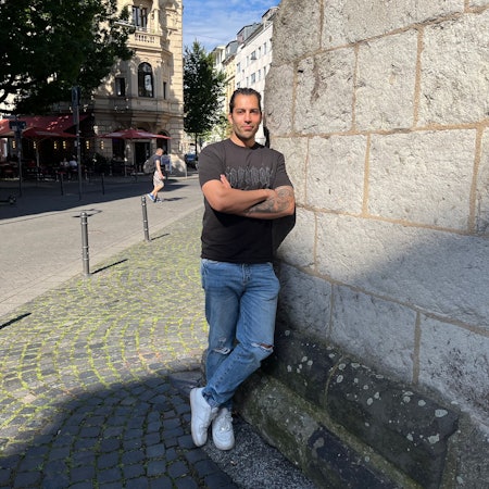 Siavash Hosseini lehnt an einer Mauer der Eigelsteintorburg. Die Arme sind verschränkt, der Blick zuversichtlich.