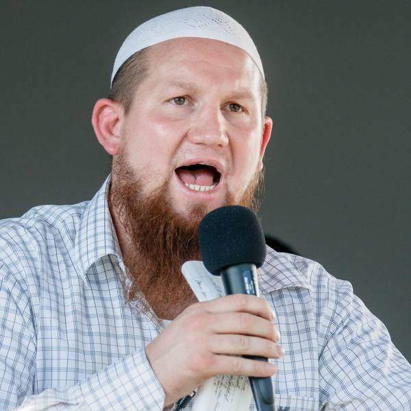 Pierre Vogel, ein weißer Mann mit längerem Vollbart und einer weißen muslimischen Gebetskopfbedeckung und kariertem Hemd. Er hält ein Mikro in der Hand und spricht zu einem Publikum.