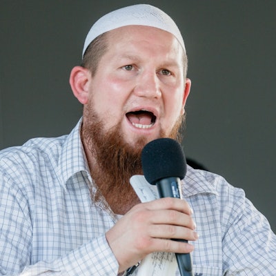 Pierre Vogel, ein weißer Mann mit längerem Vollbart und einer weißen muslimischen Gebetskopfbedeckung und kariertem Hemd. Er hält ein Mikro in der Hand und spricht zu einem Publikum.
