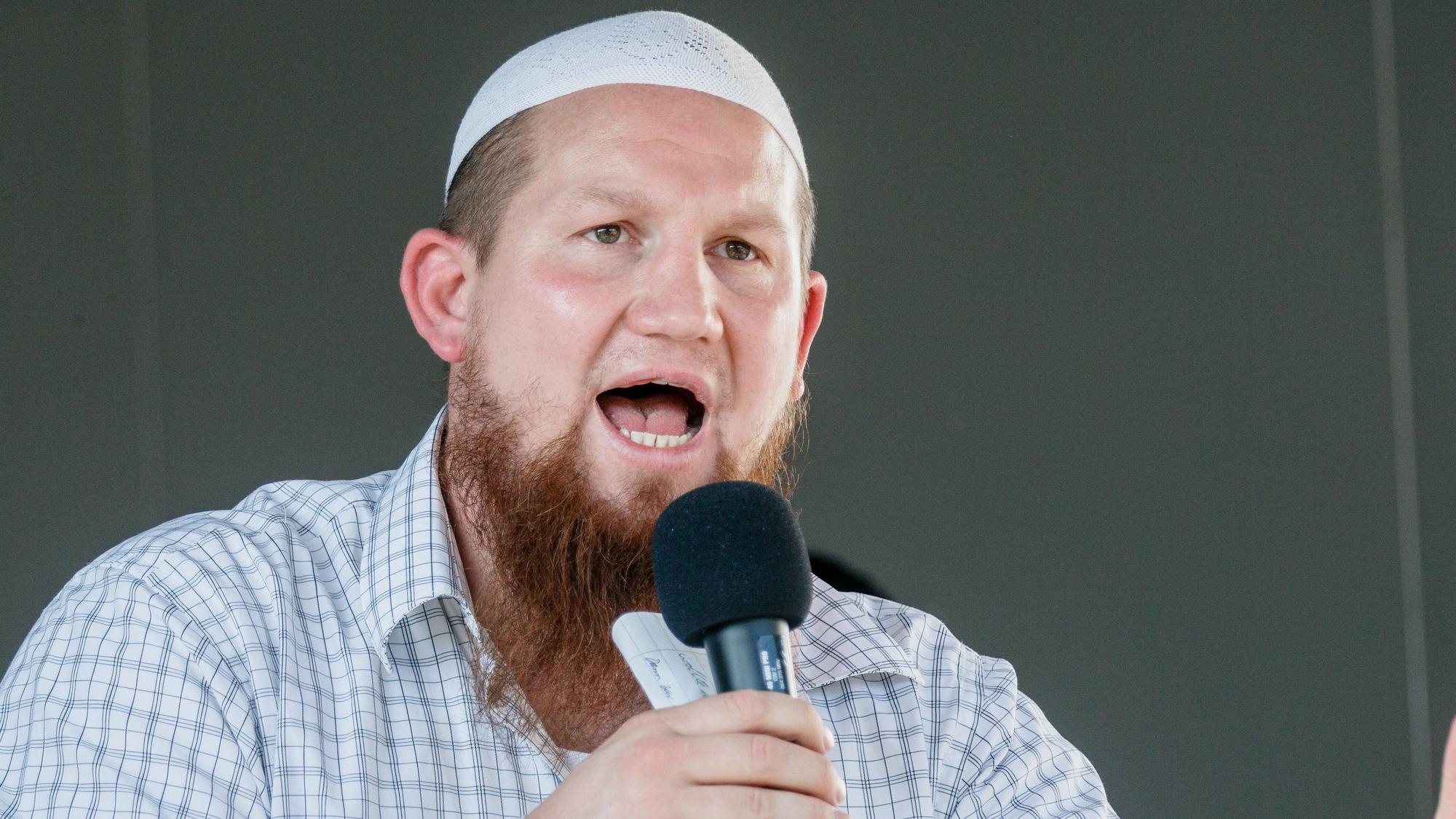 Pierre Vogel, ein weißer Mann mit längerem Vollbart und einer weißen muslimischen Gebetskopfbedeckung und kariertem Hemd. Er hält ein Mikro in der Hand und spricht zu einem Publikum.