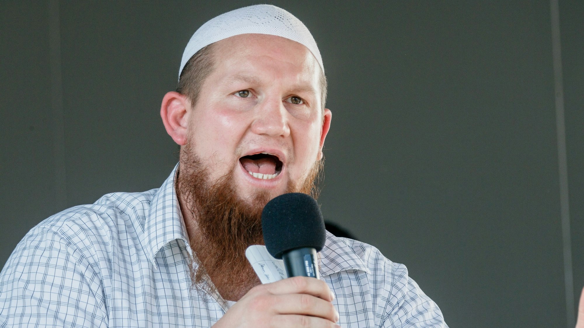 Pierre Vogel, ein weißer Mann mit längerem Vollbart und einer weißen muslimischen Gebetskopfbedeckung und kariertem Hemd. Er hält ein Mikro in der Hand und spricht zu einem Publikum.