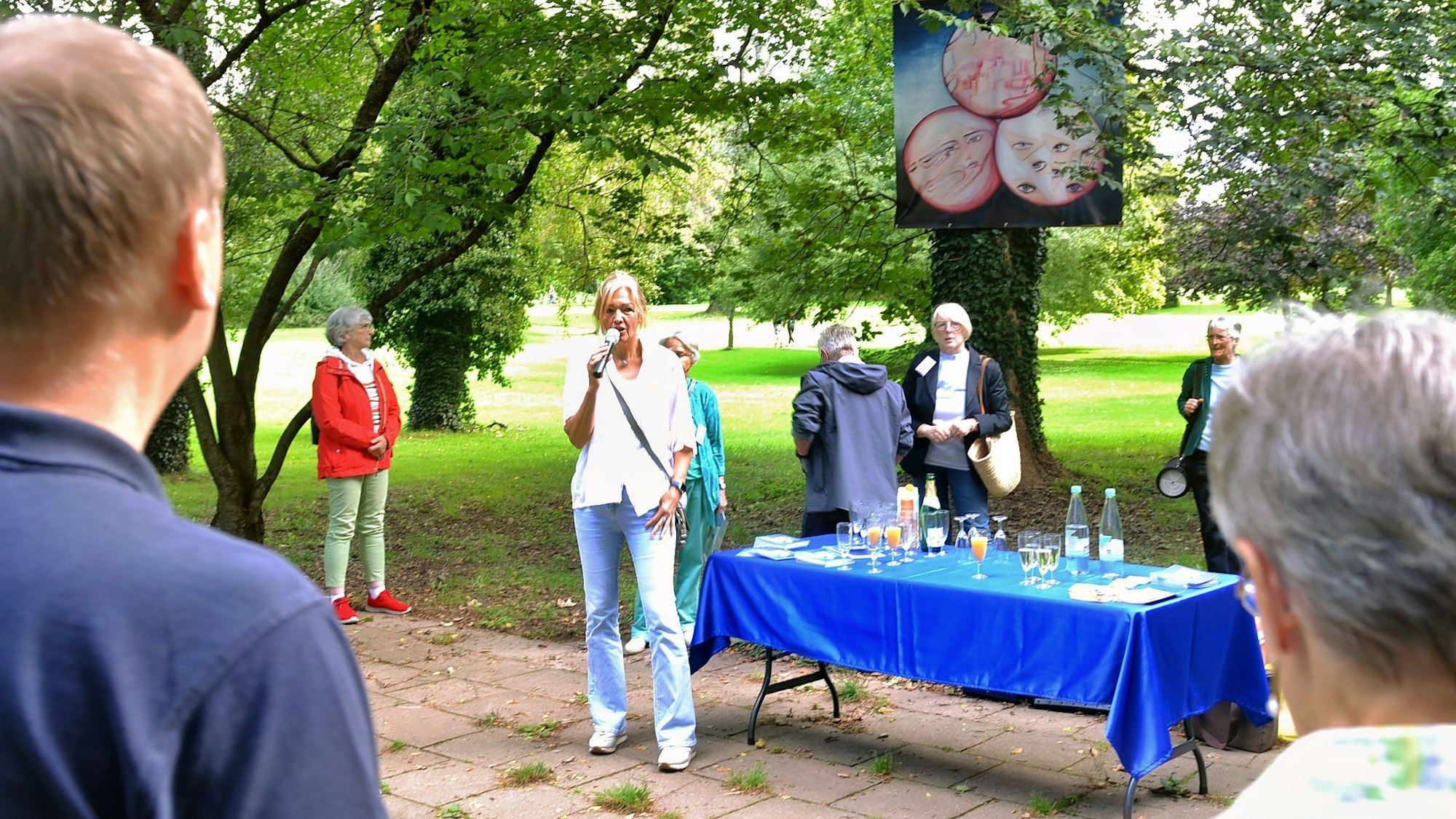 Stening spricht zum Publikum, neben ihr ein Tisch mit Getränken. Im Hintergrund hängt eines der Kunstwerke an einem Baum.