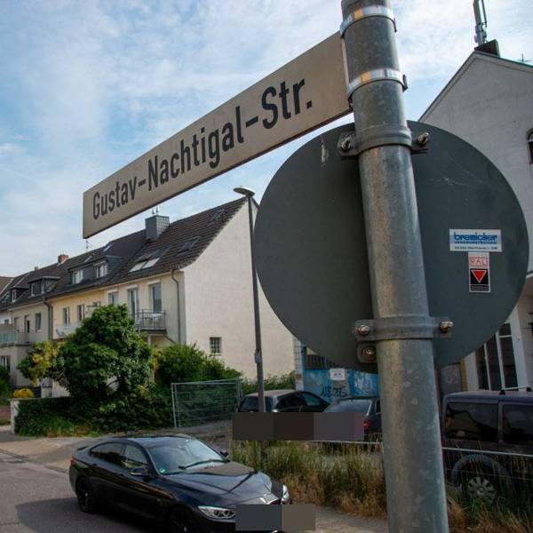 Ein Straßenschild und die dazugehörige Straße mit Reihenhäusern und parkenden Autos ist zu sehen.