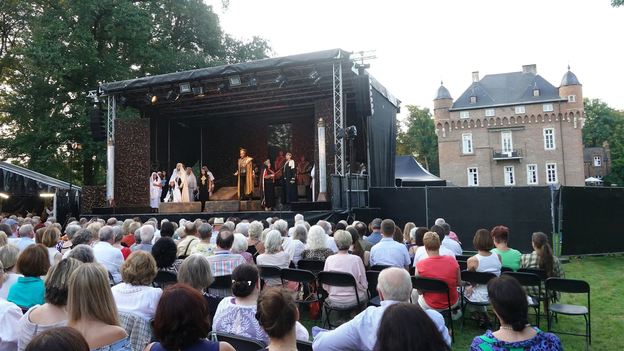 Eine kleine Menge sitzt auf Klappstühlen vor einer Bühne. Im Hintergrund das Schloss.