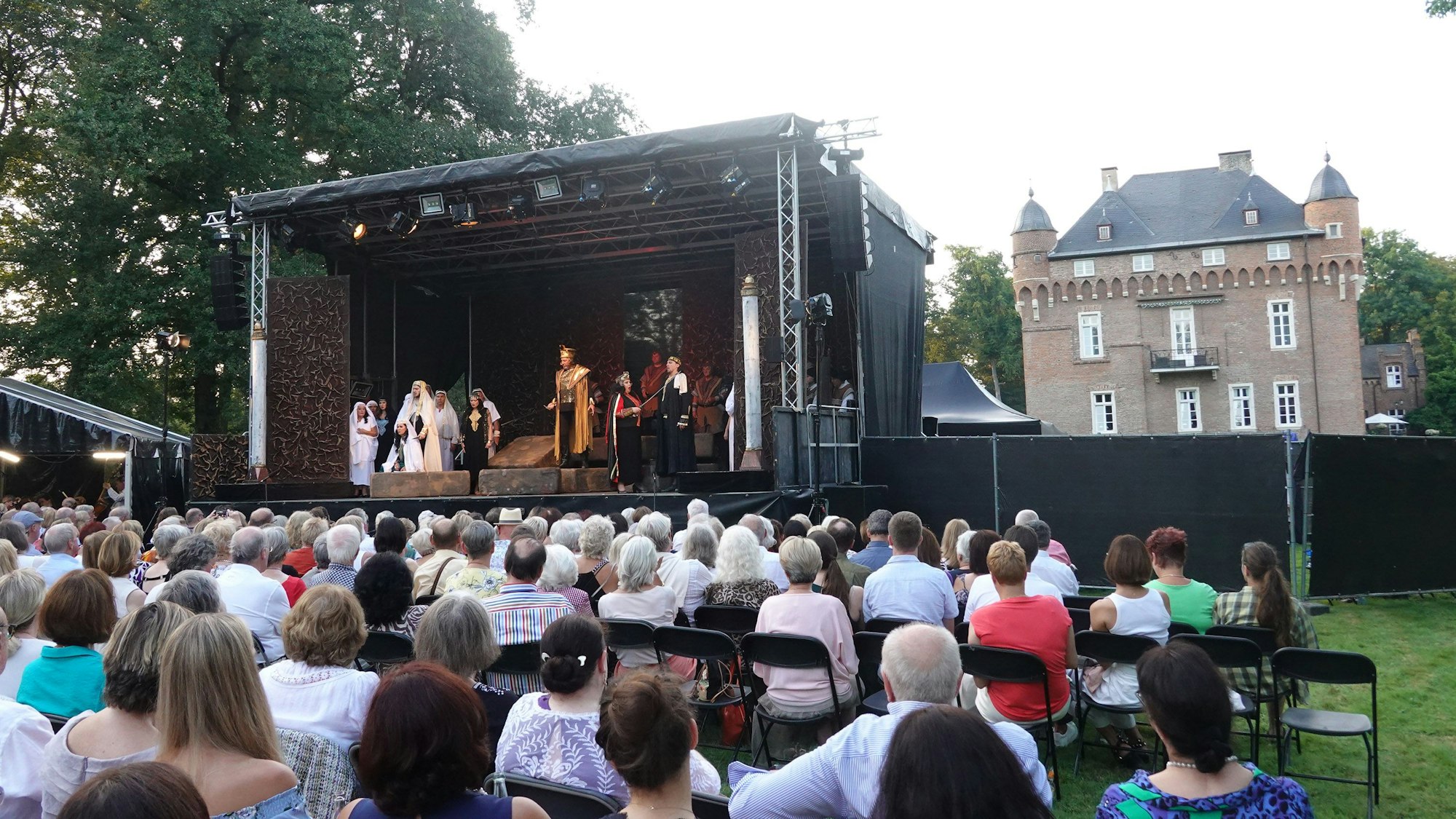 Eine kleine Menge sitzt auf Klappstühlen vor einer Bühne. Im Hintergrund das Schloss.
