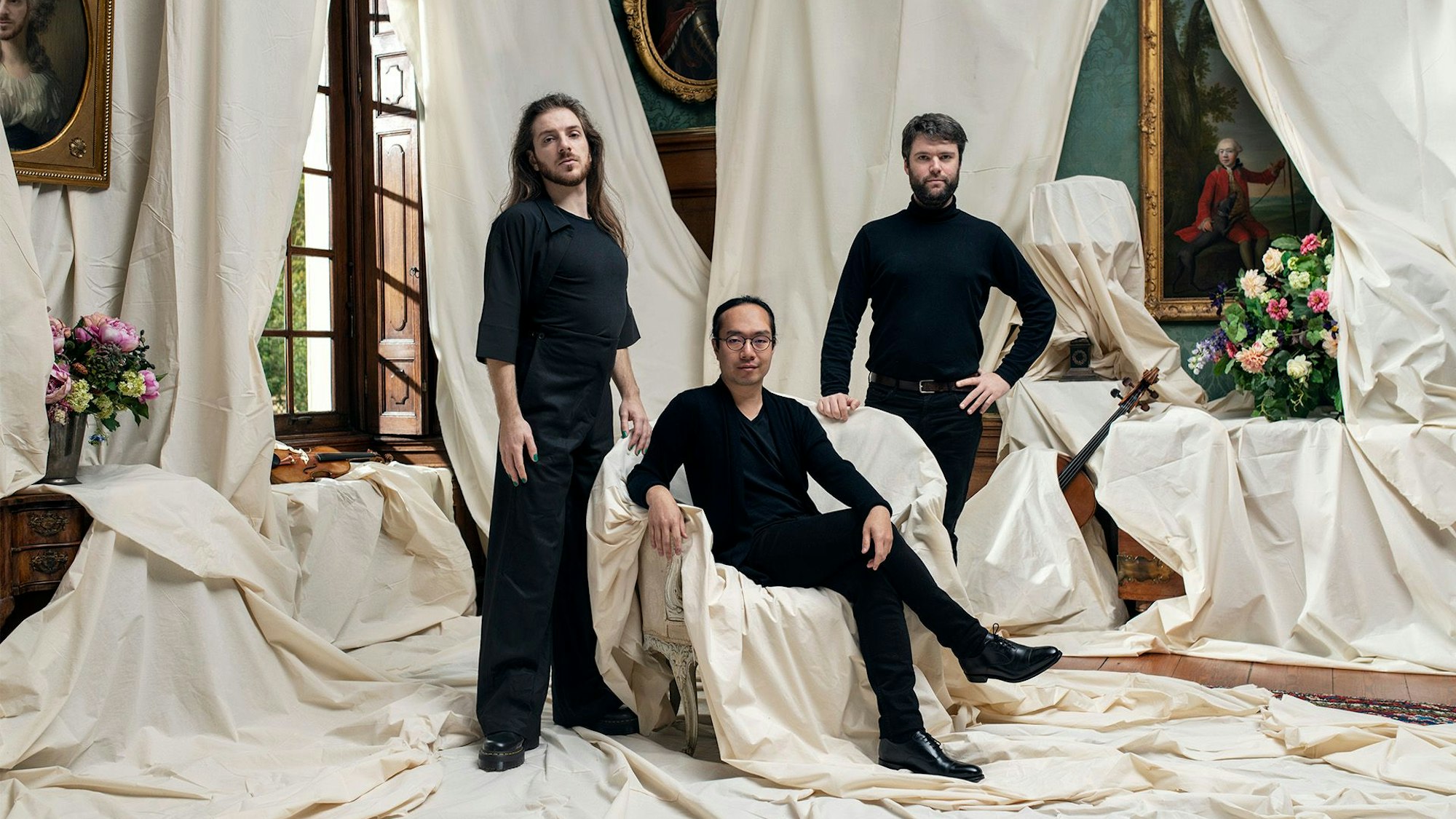 Zu sehen sind drei schwarz gekleidete Männer in einem mit Tüchern abgedeckten barocken Raum.
