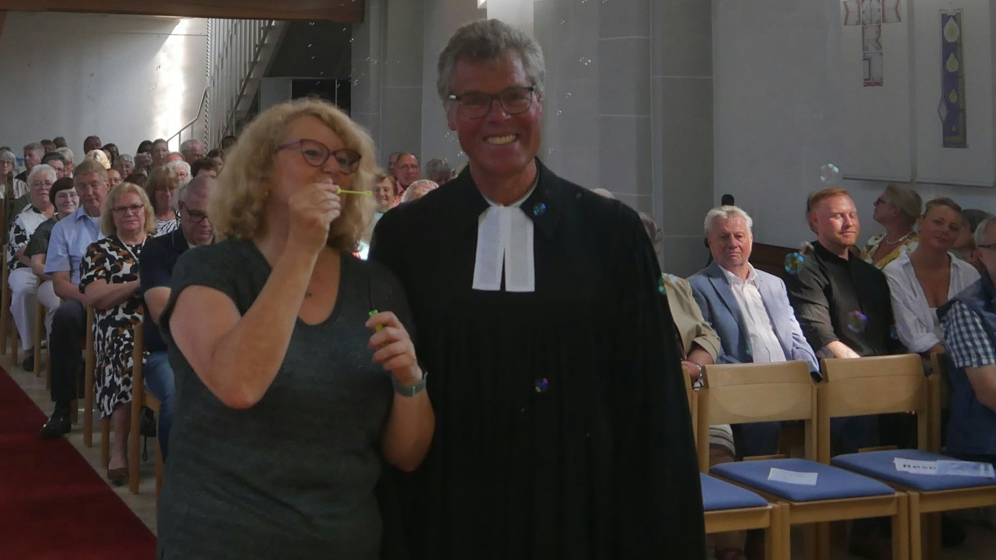 Eine Frau mit Brille und ein Pfarrer im Talar. Die Frau pustet Seifenblasen in die Kirche.