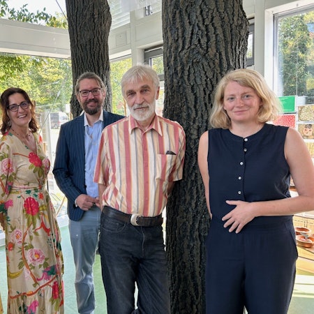 Eine Gruppe von Männern und Frauen steht zwischen Bäumen. Sie tragen sommerliche Kleidung.