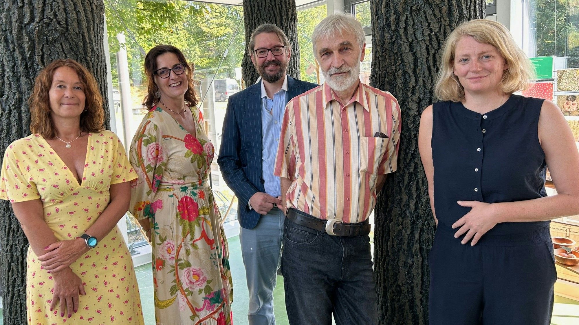 Eine Gruppe von Männern und Frauen steht zwischen Bäumen. Sie tragen sommerliche Kleidung.