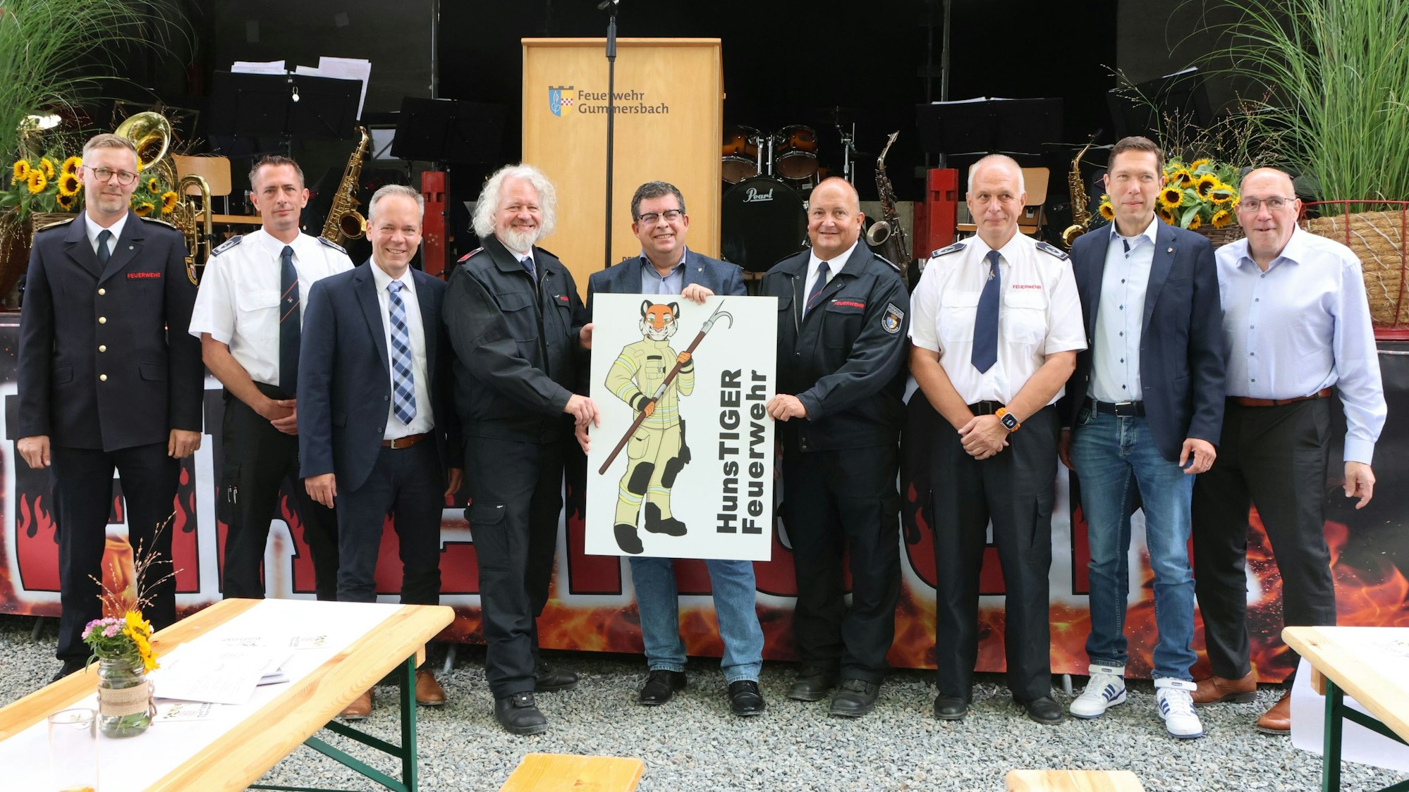 Neun Männer aus der Feuerwehr und Politik stehen mit einem Schild des Löschzugs Hunstig vor einer Bühne.
