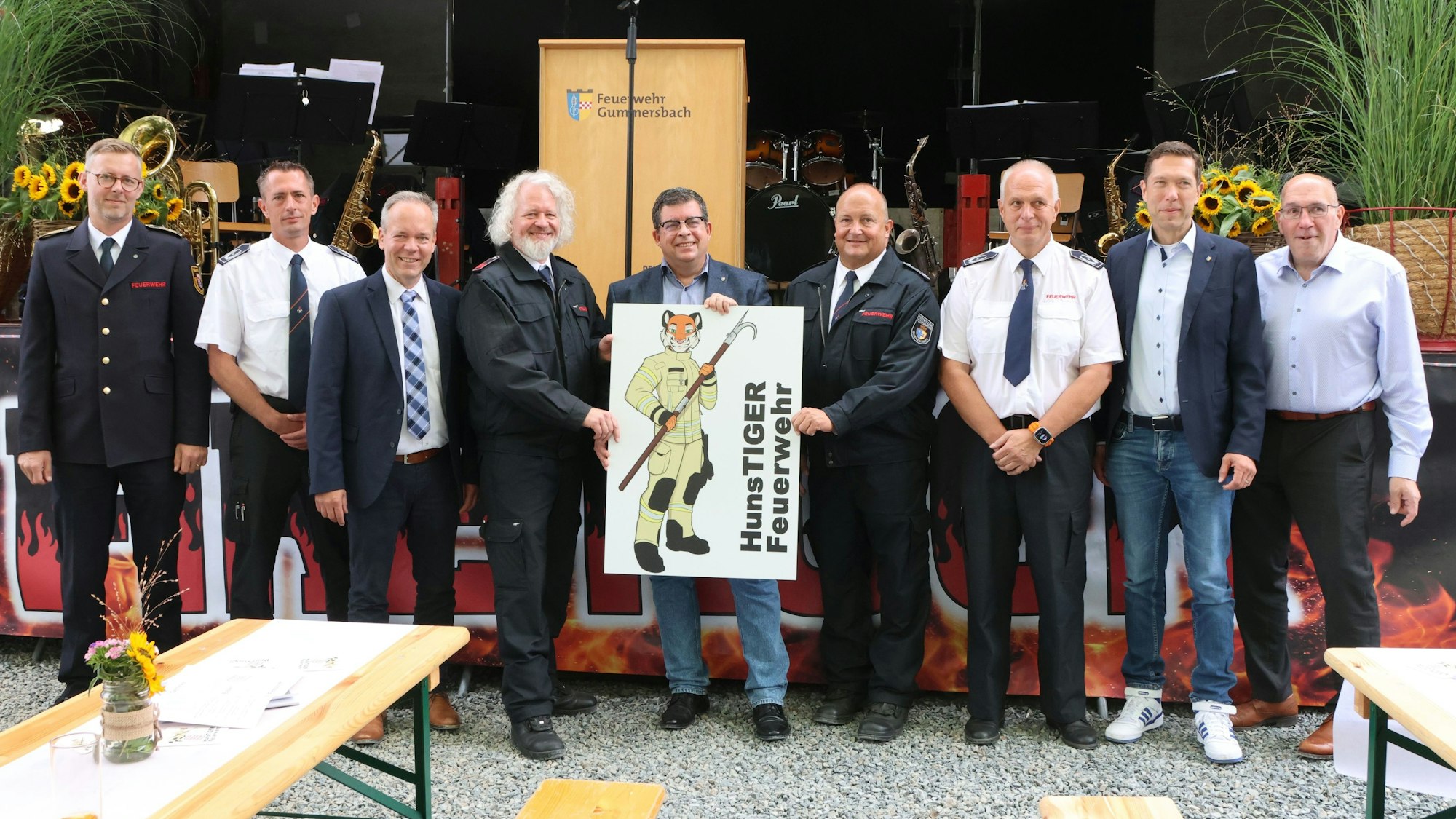 Neun Männer aus der Feuerwehr und Politik stehen mit einem Schild des Löschzugs Hunstig vor einer Bühne.