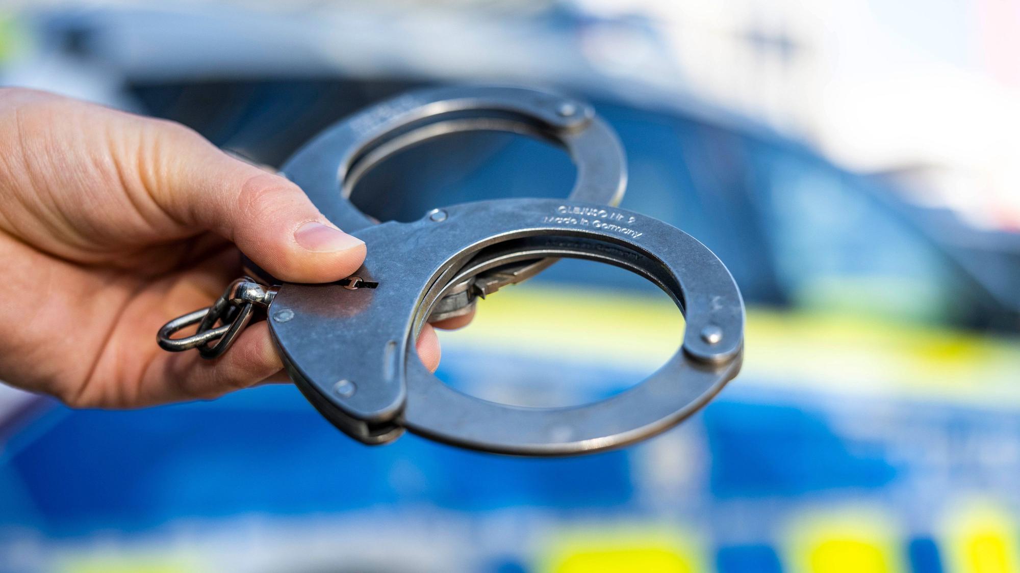 Eine Hand hält Handschellen vor einen Streifenwagen der Polizei. (Symbolbild)