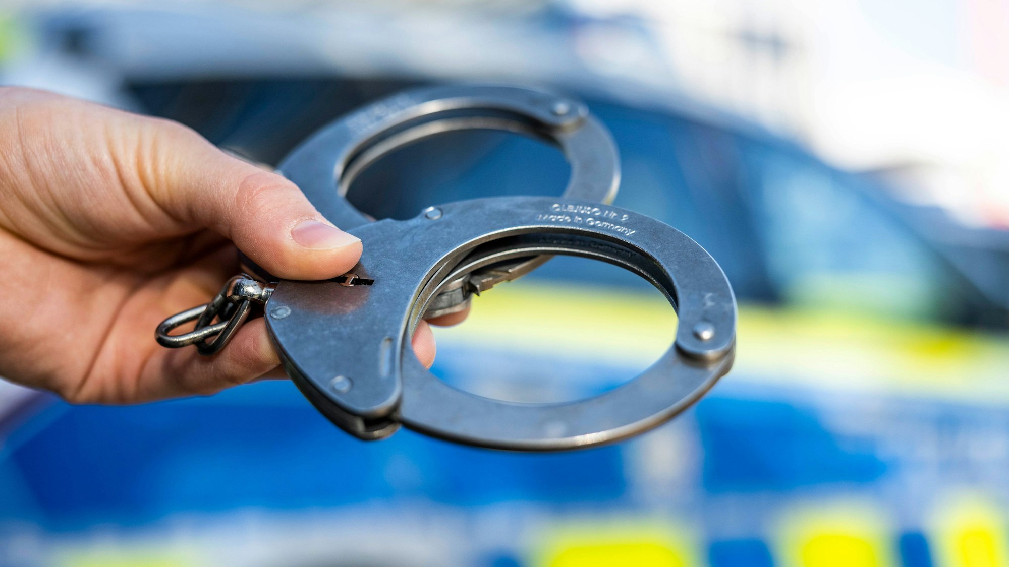 Eine Hand hält Handschellen vor einen Streifenwagen der Polizei. (Symbolbild)