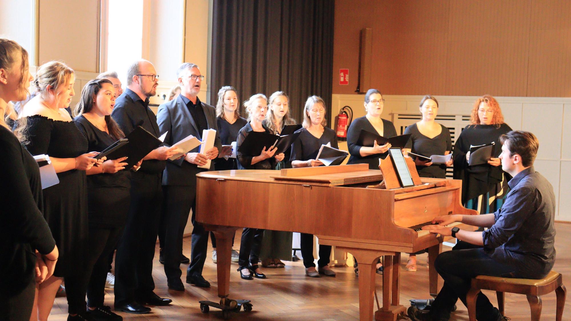 Sängerinnen und Sänger, alle in Schwarz gekleidet, haben sich um einen Flügel gruppiert, auf dem ein Pianist spielt.