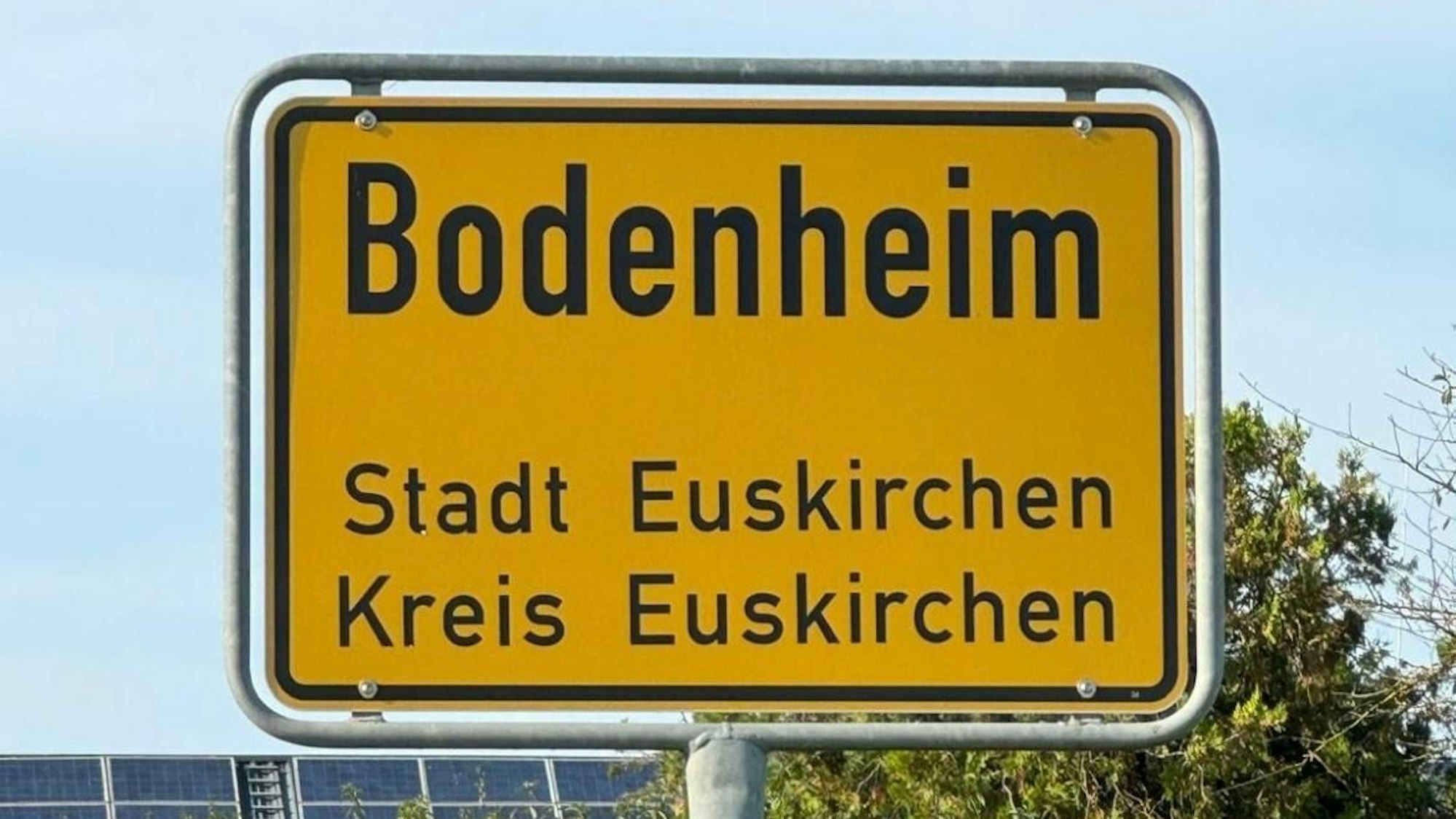 Ein gelbes Ortsschild mit der Aufschrift: „Bodenheim, Stadt Euskirchen, Kreis Euskirchen“ steht vor grünen Büschen und blauem Himmel.
