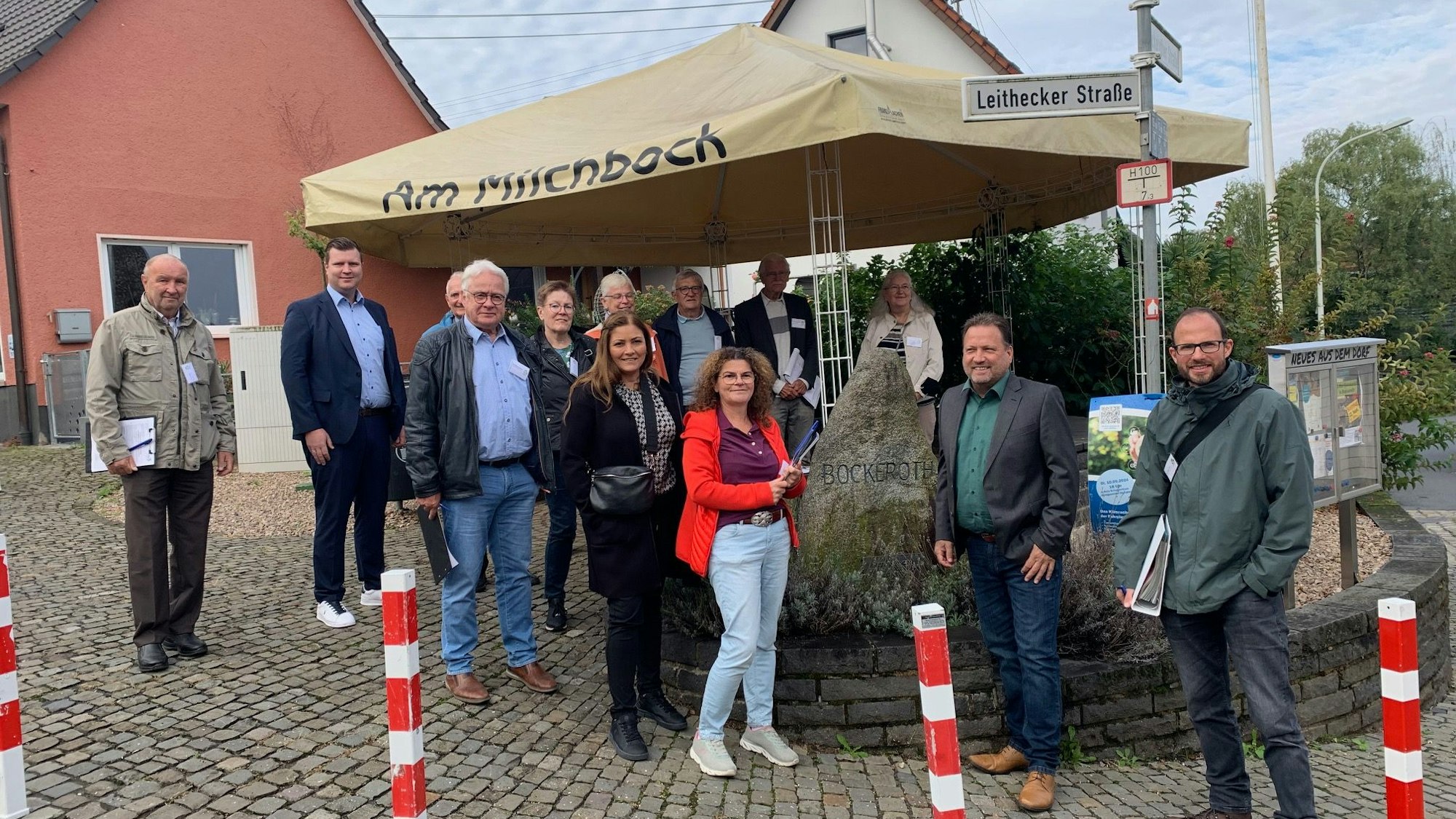 Eine Gruppe steht auf einem Dorfplatz mit Findling und Zeltdach.