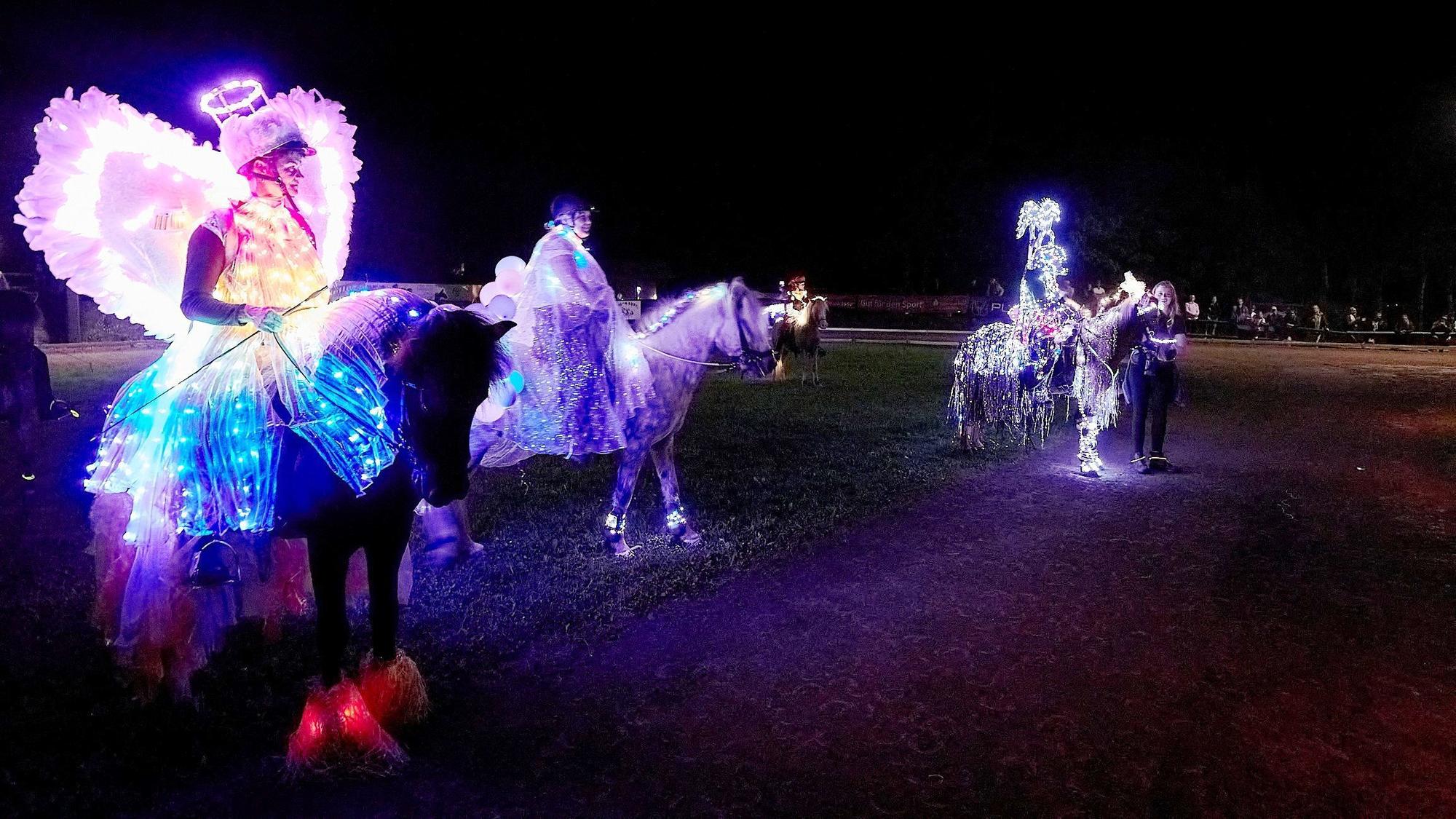 Drei Menschen sitzen in leuchtenden Kostümen auf mit Lichterketten behangenen Islandpferden in dunkler Nacht