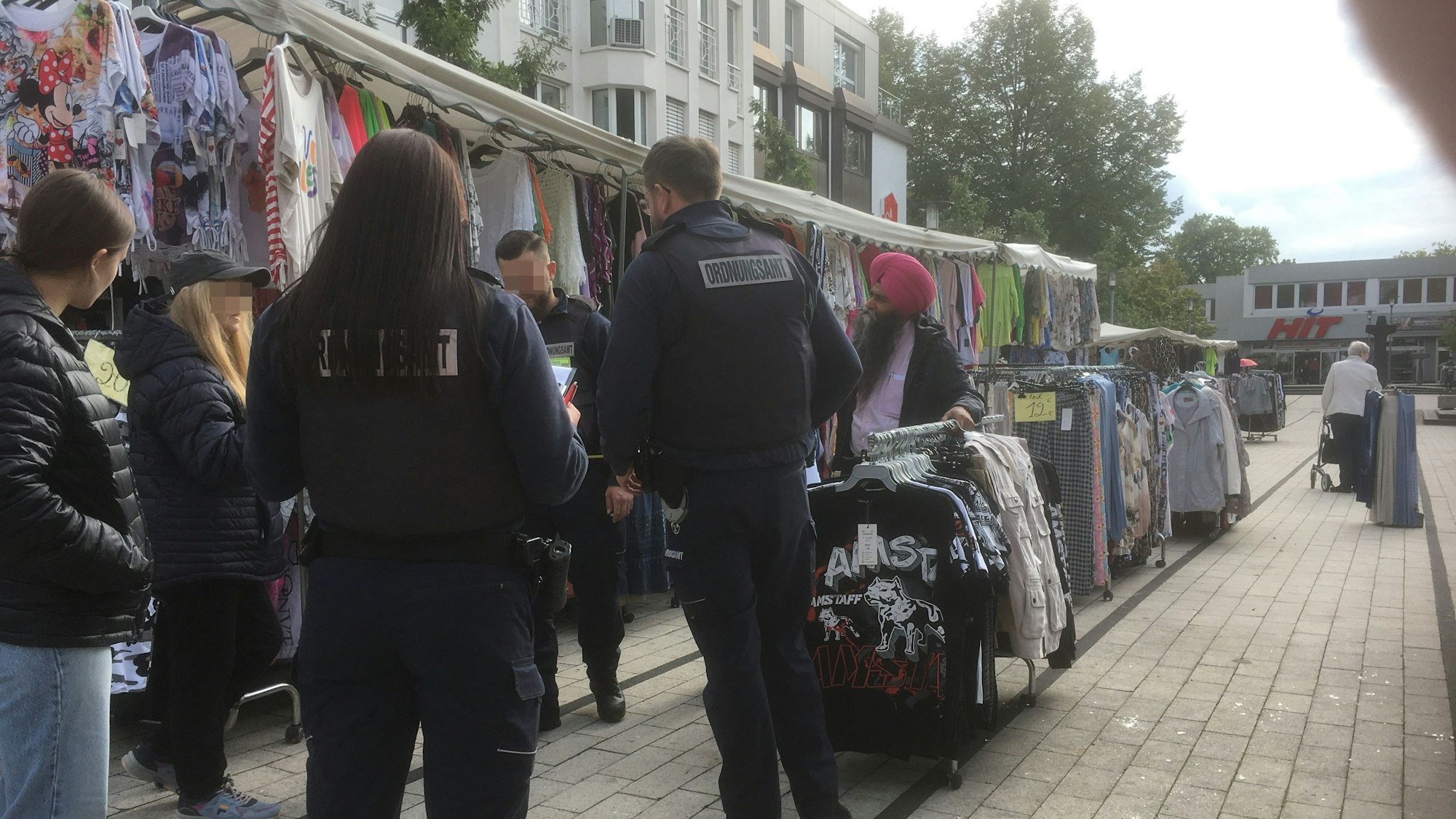 Uniformierte Mitarbeiter des Stadtordnungsdienstes an einem Marktstand mit Kleidung.
