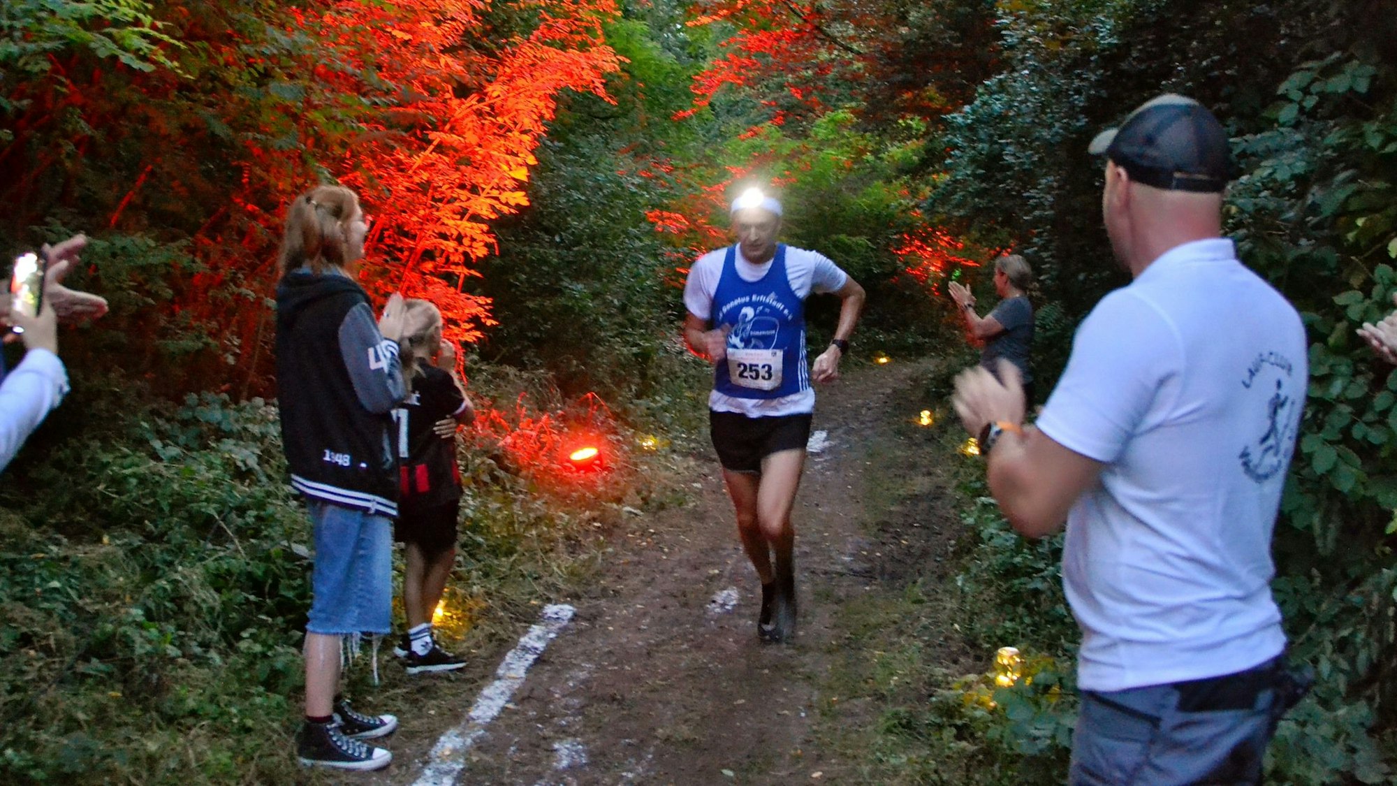 Ein Läufer mit Stirnlampe auf dem Kopf läuft über einen Waldweg. Zuschauer applaudieren ihm aufmunternd zu.