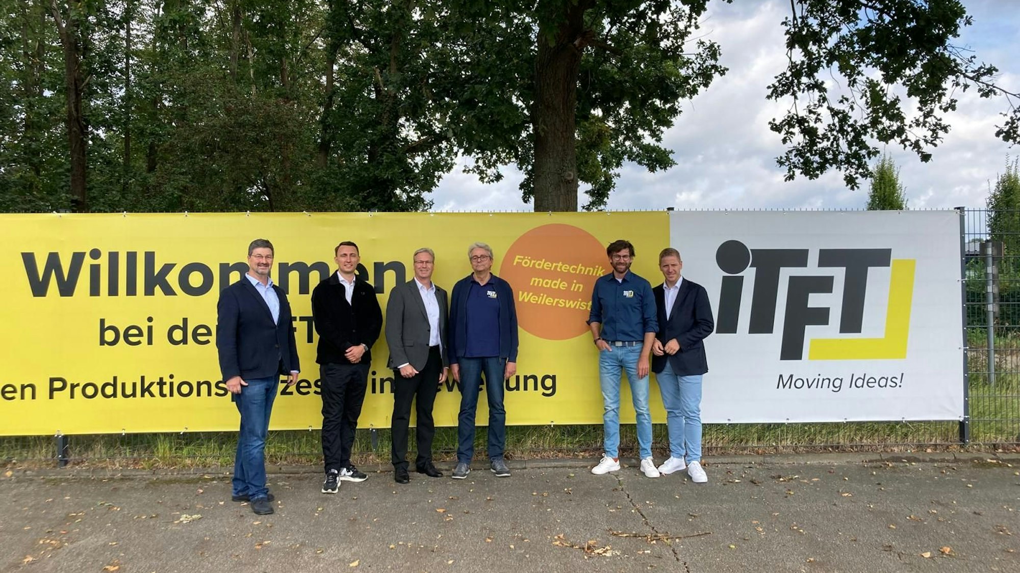 Mehrere Männer stehen vor einem Banner des Unternehmens ITFT mit der Aufschrift „Fördertechnik made in Weilerswist“.