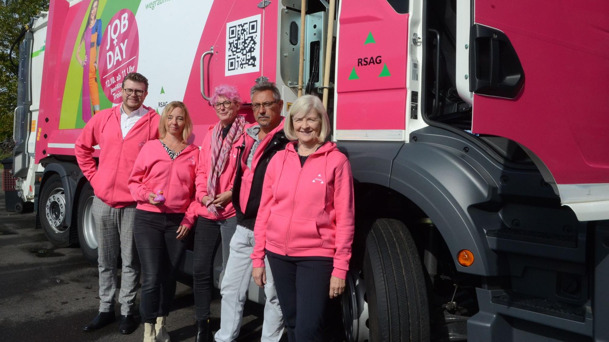 Männer und Frauen in pinkfarbenen Kapuzenjacken stehen vor einem Müllfahrzeug, das ebenfalls zu Teilen in Pink foliert ist.