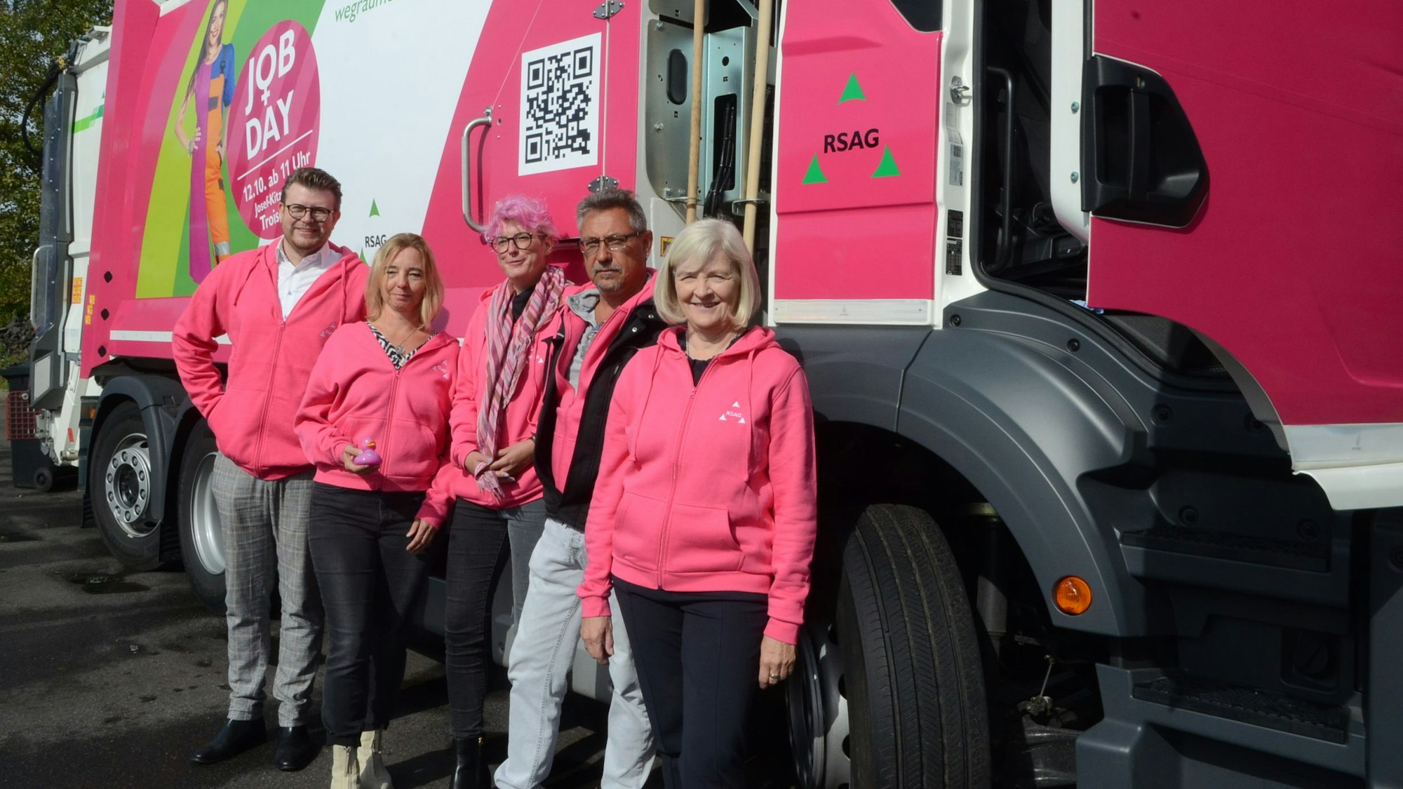 Männer und Frauen in pinkfarbenen Kapuzenjacken stehen vor einem Müllfahrzeug, das ebenfalls zu Teilen in Pink foliert ist.