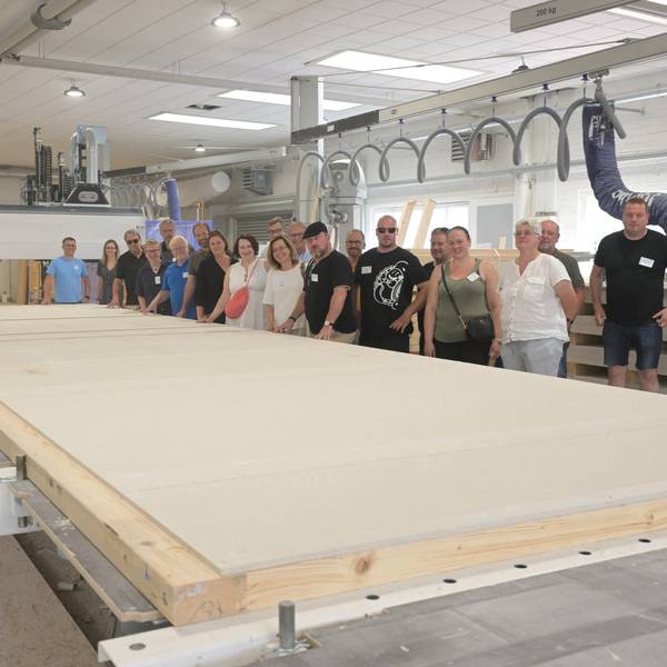Eine Gruppe von Menschen steht in einer großen Fertigungshalle. Vor ihnen liegt ein große Holzplatte, die einmal eine Hauswand werden soll.