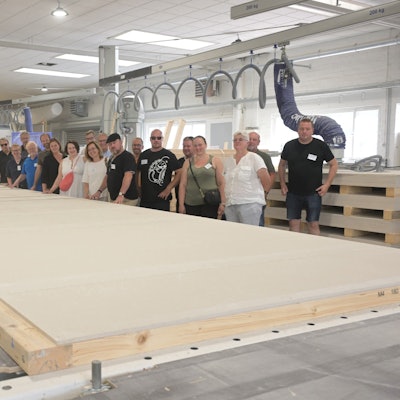 Eine Gruppe von Menschen steht in einer großen Fertigungshalle. Vor ihnen liegt ein große Holzplatte, die einmal eine Hauswand werden soll.