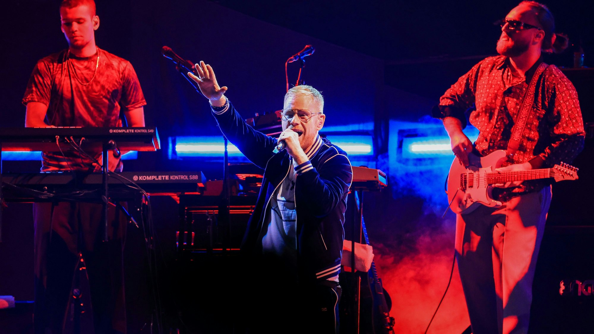 Peter Fox steht singend mit erhobenem Arm auf der Bühne in der Kölner Lanxess-Arena, hinter ihm ein Keyboarder und ein Gitarrist.