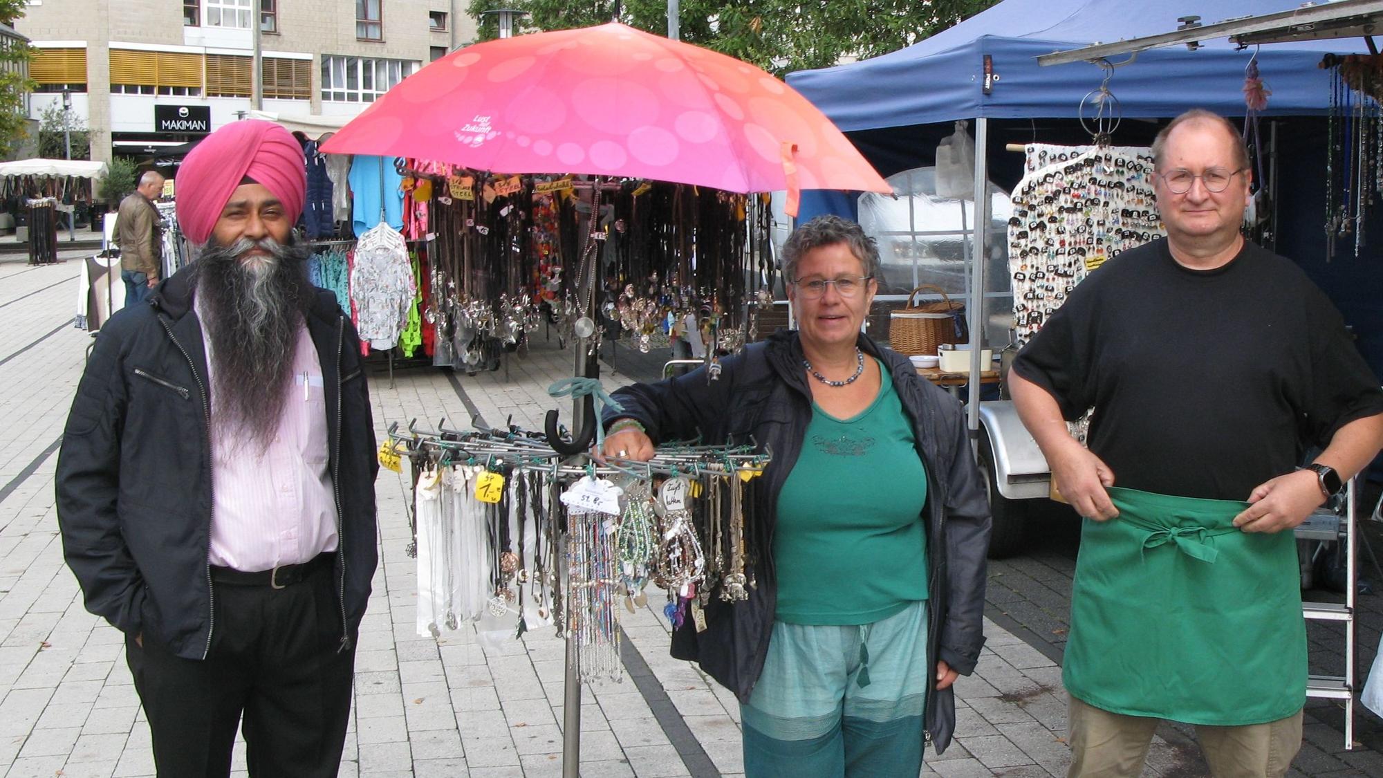 Zwei Markthändler und eine Markthändlerin stehen an einem Schmuckstand auf dem Marktplatz.