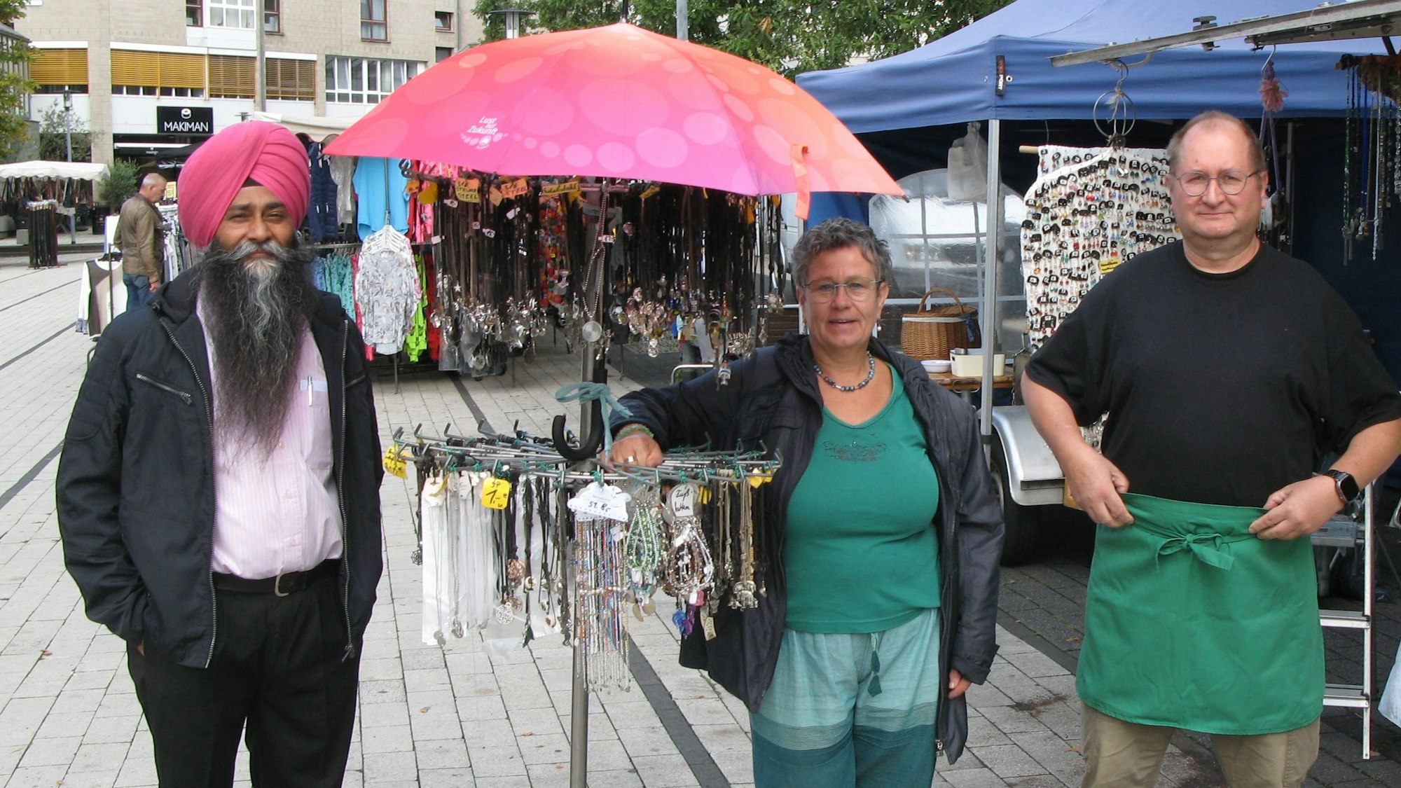 Zwei Markthändler und eine Markthändlerin stehen an einem Schmuckstand auf dem Marktplatz.