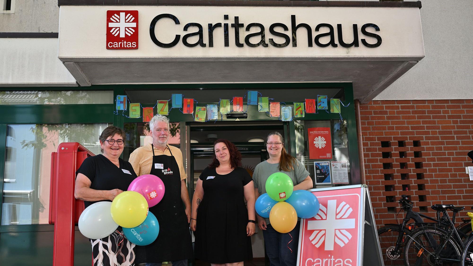Drei Frauen und ein Mann stehen vor einem Gebäude mit der Aufschrift Caritashaus. Sie halten bunte Luftballons in der Hand.