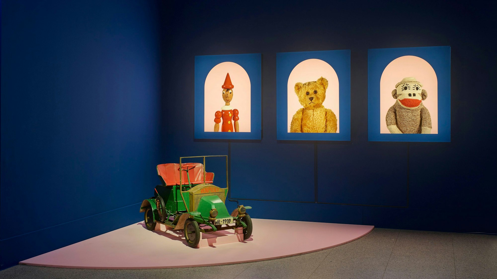 Ein Holzauto steht vor Porträts von Stofftieren und einem Harlekin.