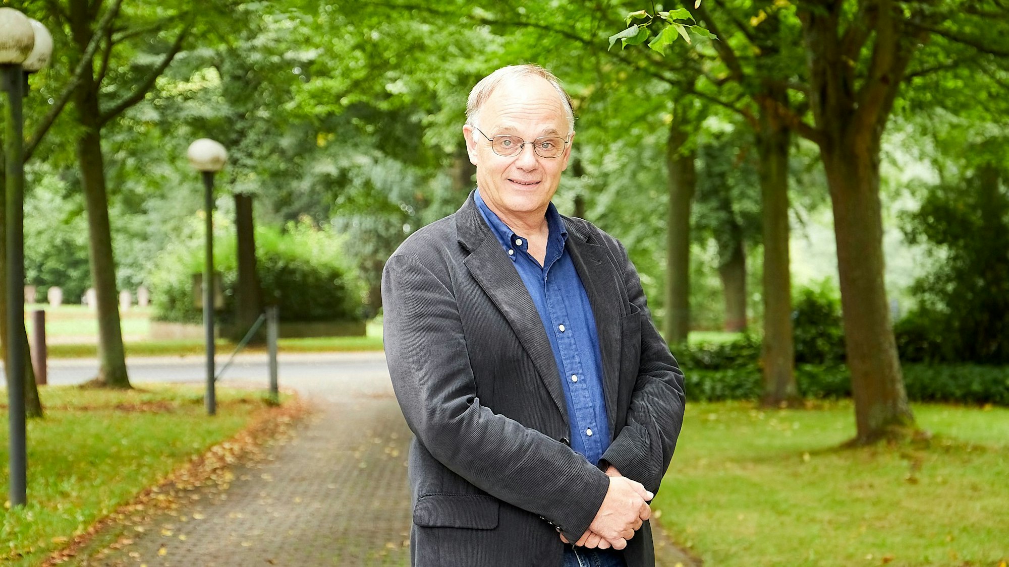 Ein Herr mit Brille legt in einem Park die Hände vor dem Bauch übereinander.