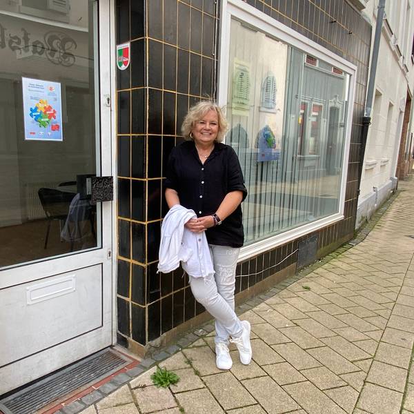 Eine Frau in schwarzer Bluse und heller Hose lehnt an einer schwarz gekachelten Hauswand.