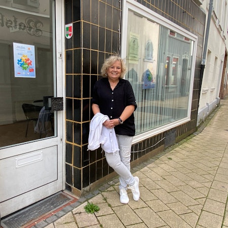 Eine Frau in schwarzer Bluse und heller Hose lehnt an einer schwarz gekachelten Hauswand.