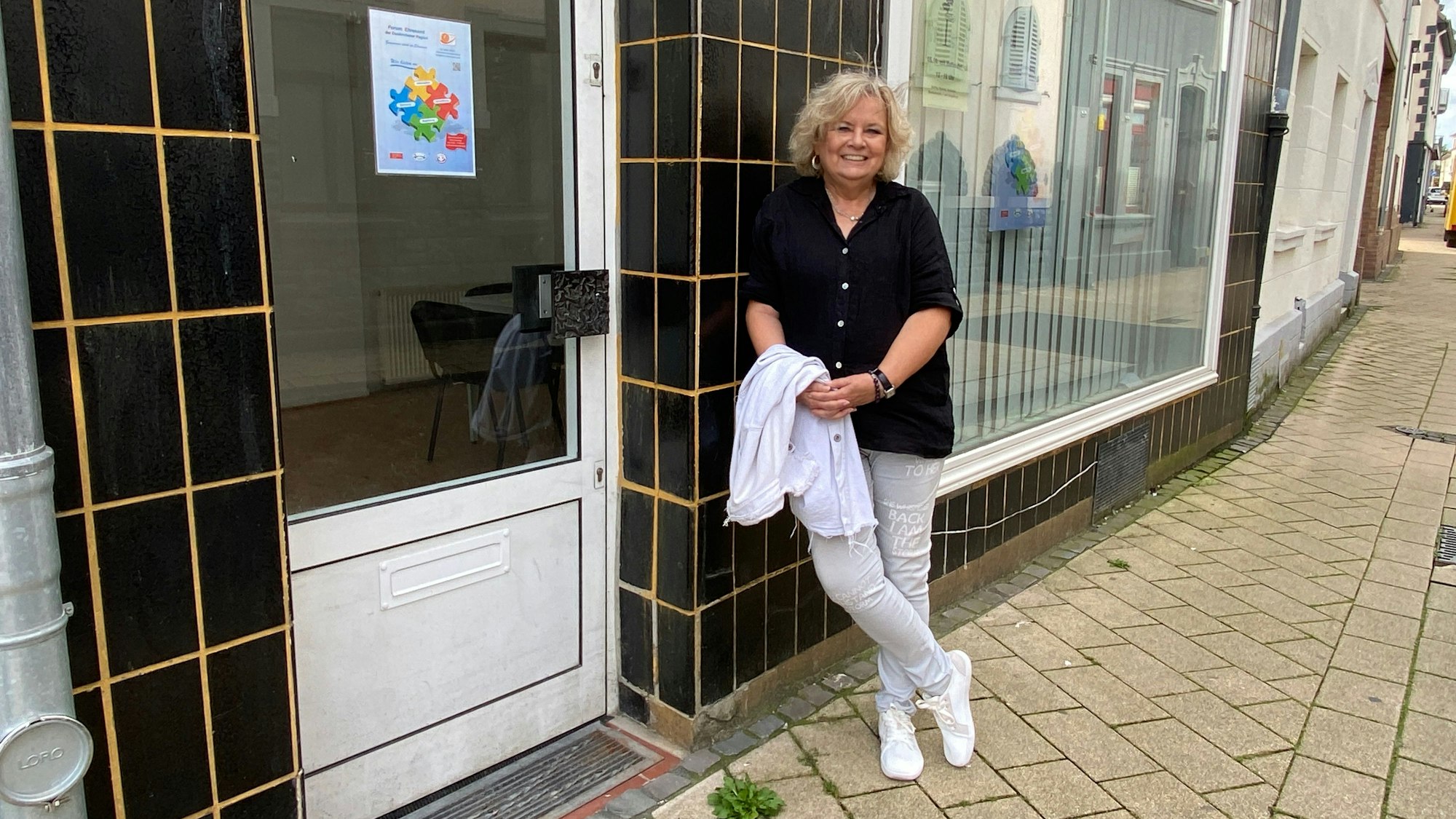 Eine Frau in schwarzer Bluse und heller Hose lehnt an einer schwarz gekachelten Hauswand.