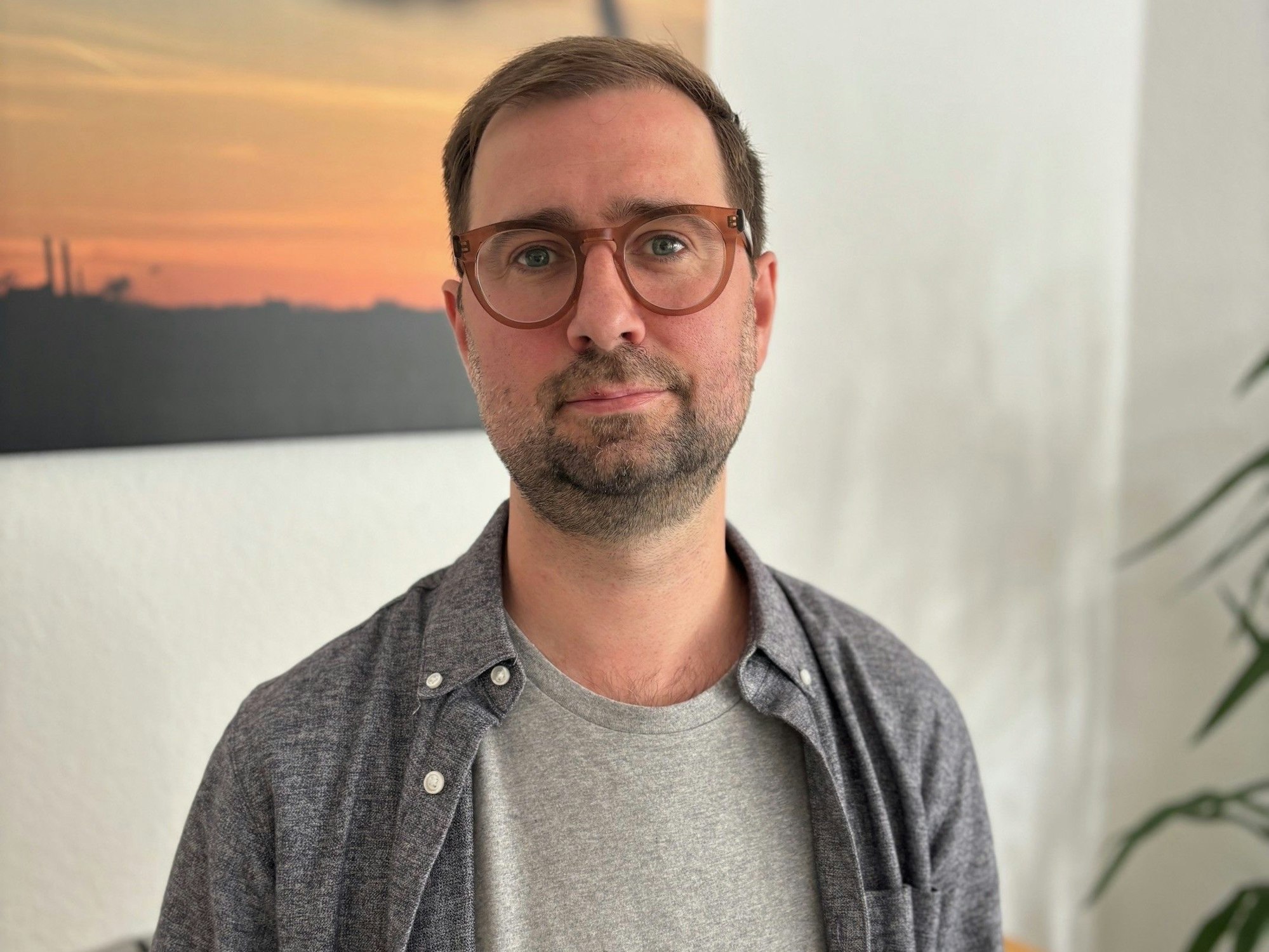 Auf dem Bild ist ein Portrait von Dominik Schmitz zu sehen. Er trägt ein graues T-Shirt, eine Brille und ein graues Hemd.